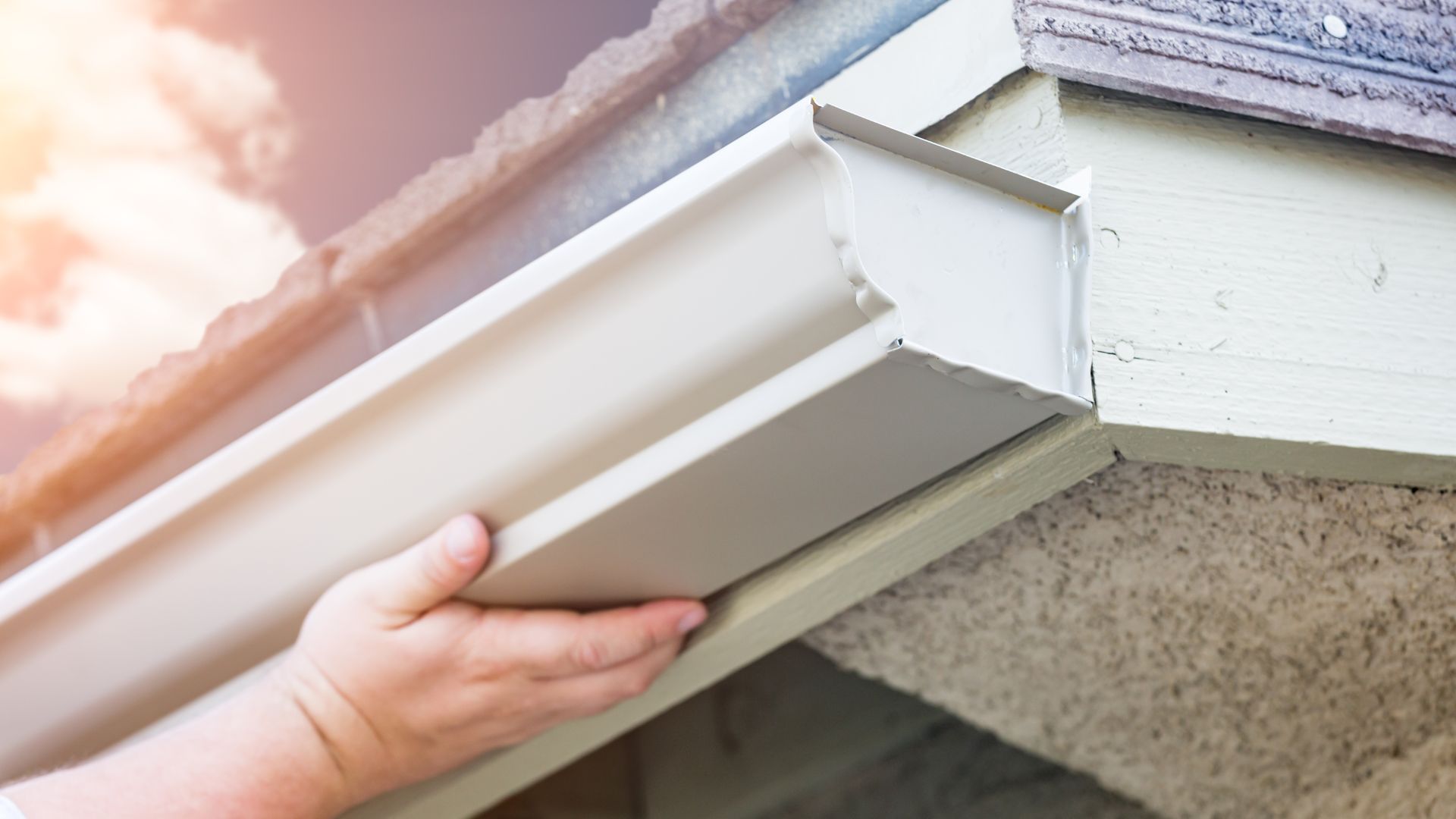 Hand installing white gutter along roof edge of building